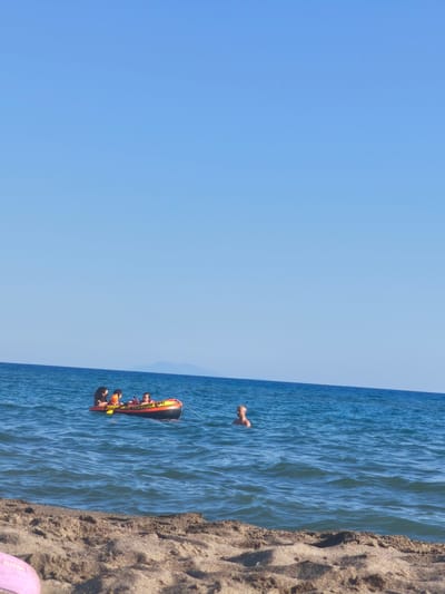 In the Tyrrhenian Sea off the coast of Castiglione della Pescaia, Italy, on a clear, sunny day, a bright orange and yellow inflatable tube is being towed by an unseen boat. Four individuals, appearing to be a family, are riding on the tube. A man with short, dark hair is in the front, steering or holding on. Behind him, a woman with dark hair is seated, also holding on. Two children are positioned further back on the tube. To the right of the inflatable tube, a man with light-colored hair is swimming in the sea, facing away from the camera. The water is a vibrant blue, with small waves rolling towards the sandy beach in the foreground. The beach is covered in fine, light-brown sand, with some debris and footprints visible. In the distance, a faint, hazy outline of a mountainous island can be seen on the horizon under a clear, pale blue sky. The overall scene depicts a leisurely day at the beach, with water sports and swimming.