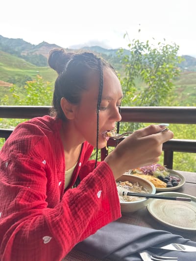 An adult woman is depicted in a side profile, seated outdoors on what appears to be a balcony or terrace, in Púng Luông, Vietnam. She is positioned to the left of the frame, facing right, with her eyes downcast towards a spoonful of food she is bringing to her mouth. Her dark hair is styled in a high bun, with several thin braids framing her face, some adorned with small silver, star-shaped embellishments. She wears a red, textured, long-sleeved garment featuring embroidered white heart motifs, over a light yellow-green undergarment. Her right hand, with long, pointed nails painted with a greenish-blue iridescent polish, holds a silver spoon from which she is consuming a portion of food.
On a dark, woven table in front of her, a meal is laid out. This includes a light-colored bowl containing noodles or soup with dark chopsticks resting on its rim, and a grey/beige plate holding a salad composed of purple cabbage, lettuce, and red onions, alongside what appears to be an omelet or frittata and several dark olives or berries. An additional empty grey/beige plate is also present. A dark grey folded fabric napkin, holding a silver fork and knife, is situated in the foreground.
The background reveals a panoramic view of rolling green mountains, consistent with terraced agricultural landscapes common in Púng Luông. Lush green foliage and trees are visible immediately beyond a dark metal railing, which delineates the edge of the terrace. The sky overhead is bright and appears overcast.