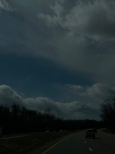 A dimly lit photograph shows a highway running to the right of the frame. A vehicle is present on the road. Alongside the highway, a dark, shadowed area filled with trees and greenery is visible. The sky above is filled with clouds, some dark and overcast, adding to the somber atmosphere of the image. This photograph was taken near the Town of Fishkill, United States of America.