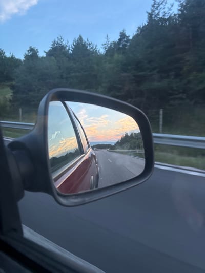 The image shows a rearview mirror reflection of a highway at dusk. The reflection displays a road with lane markings, a guardrail, and trees lining the side of the road. The sky in the reflection is filled with colorful clouds, suggesting a sunset. The original image captures a side view of a car, with the rearview mirror prominently in the foreground, showing the reflected scenery. The location is identified as Baliovtsi, Bulgaria.