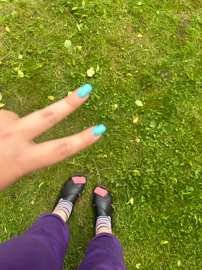 The image shows a pair of legs and feet from a person's perspective, sitting on grass. The person is wearing purple pants and black slip-on sandals with pink accents. They are also wearing socks with horizontal stripes in pink, white, and blue. One hand is visible, making a peace sign with brightly painted turquoise fingernails. The background is a grassy lawn with some fallen leaves, suggesting an outdoor setting. The lighting indicates it is daytime, likely a clear day given the sharp shadows. There is no visible text in the image. The overall mood appears casual and relaxed, with the person possibly enjoying a moment outdoors in Leningradskaya, Russia.