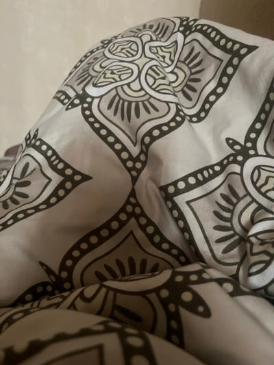 The image shows a close-up of a fabric with a mandala-like pattern. The fabric is draped and appears to be part of bedding or upholstery. The pattern consists of intricate geometric shapes and floral elements in shades of grey, white, and black. The lighting is soft, and the focus is on the texture and design of the fabric. There is no discernible location in the city of Vladikavkaz, Russia, or any identifiable people, actions, or interactions. The image is purely a study of the textile's pattern and drape.