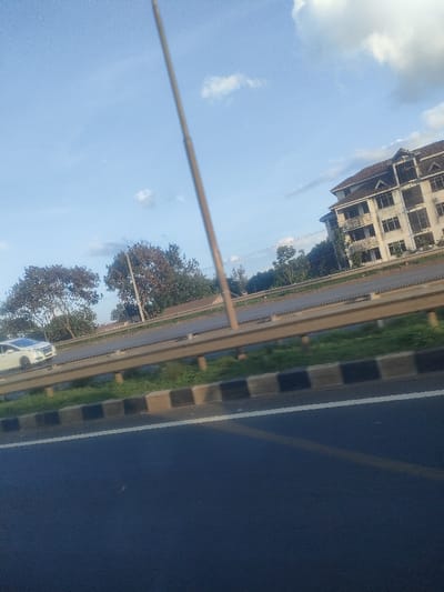 A road scene in Juja ward, Kenya, captured from a moving vehicle, displays a section of asphalt road in the foreground with a faded yellow lane line and a distinct white edge line. This road borders a black and white striped curb, which gives way to a grassy verge. Behind the verge, a metallic guardrail runs horizontally across the mid-ground. Beyond the guardrail, a multi-lane highway is visible, on which a white sedan or small SUV travels in the opposite direction on the leftmost lane. A tall, slender, light-colored pole extends diagonally from the bottom-center to the upper-center of the frame, partially obscuring the view. Clusters of green trees are present on the far side of the highway. In the background, on the right side, a multi-story building with a prominent dark, sloped roof and multiple windows is partially visible. The sky overhead is clear blue with scattered white cumulus clouds. No individuals are depicted.