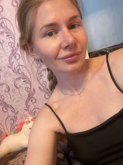 A young woman with blonde hair in a braid is taking a selfie. She is wearing a black tank top and a silver necklace. To her left, a unicorn plush toy is visible. The background consists of pink patterned wallpaper and a blue wall. The image appears to be taken indoors, possibly in a residential setting. There is no information to indicate the location is Маринопіль, Ukraine.