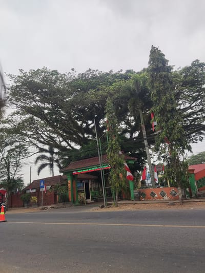 The image captures the entrance of SMA Negeri 1 Buluagung, a public high school in Buluagung, Indonesia. The school gate is decorated with green and orange accents, and red and white Indonesian flags are displayed prominently. The school's welcome message is visible on a digital signboard. Towering trees, including a large, mature tree with sprawling branches and a tall, slender tree, dominate the background, suggesting a lush, green environment. A paved road runs in front of the school, with a pedestrian crossing sign visible on the sidewalk. The sky is overcast, indicating a cloudy day. The scene appears calm and orderly, with no people immediately visible. The overall impression is one of a well-established educational institution set within a natural landscape.