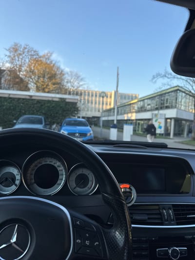 The image provides a driver's perspective from within a black Mercedes-Benz vehicle in the city of Bühl, Germany, looking forward through the windshield on a clear, sunny day.
**Vehicle Interior:** The lower portion of the image shows the vehicle's black multi-function steering wheel with the Mercedes-Benz logo. Behind the steering wheel is a three-dial instrument cluster, with a central speedometer, a tachometer to the right, and a fuel/temperature gauge to the left. The dashboard is black, featuring a central infotainment screen that appears off, and air conditioning vents. An orange-lit accessory is visible clipped to one of the central air vents.
**Exterior Scene:** The view outside is under a bright, clear blue sky. In the immediate foreground, a dark green, trimmed hedge runs horizontally. Behind the hedge, two cars are parked: a light-colored sedan is partially visible on the left, and a blue Mercedes-Benz sedan is parked further to the right. A white vertical pole, possibly a parking barrier or signpost, stands in the mid-ground, alongside a smaller white kiosk-like structure. A solitary person is visible in the mid-ground, walking from right to left across the scene, wearing a dark jacket and light-colored trousers/pants.
**Background Environment:** The background features multiple buildings and trees. On the left, a large, multi-story building with numerous windows and a distinct circular clock or emblem on its upper facade is illuminated by the sun. To its right, another modern building with extensive glass paneling and a flat roof is visible. Deciduous trees, some with remaining autumn foliage and others bare, are interspersed throughout the background. The ground appears paved, consistent with a street or parking area.