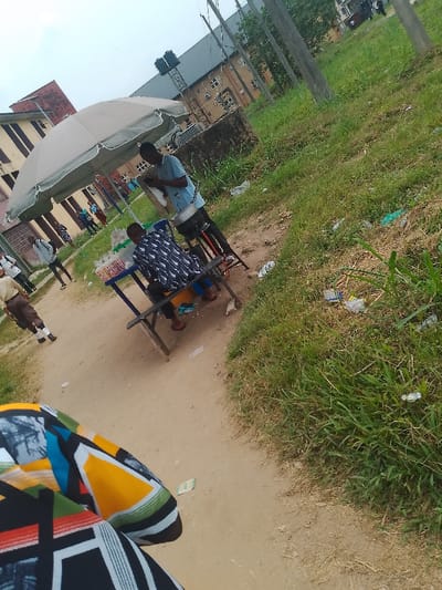 The scene is an outdoor market area in Choba, Nigeria, captured during daylight under an overcast sky. A light-colored market umbrella shades a small, unpainted wooden stall comprising a table and benches. A male vendor, wearing a light blue short-sleeved shirt and dark trousers, stands behind the table, looking down at a white object he holds. A metallic pot on a black stand is positioned to his right. A second male individual, wearing a dark blue shirt with a white pattern, is seated on a bench at the stall, with red sandals on his feet. Several small, white-wrapped items are displayed on the wooden table. The stall is situated on a dirt path that leads towards buildings in the background. To the immediate right of the path is an elevated area of unkempt grass, littered with plastic wrappers and debris. In the background, several structures are visible, including a multi-story building with reddish and beige facades and a larger building with a dark roof and multiple windows. A metal structure supporting two dark water tanks stands behind the stall. Utility poles with wires are present towards the right side of the frame. Multiple individuals are seen in the distance near the buildings and on adjacent pathways. The bottom-left foreground reveals a partial view of a person wearing a multi-colored, geometrically patterned garment, with a light green currency bill visible on the dirt path.