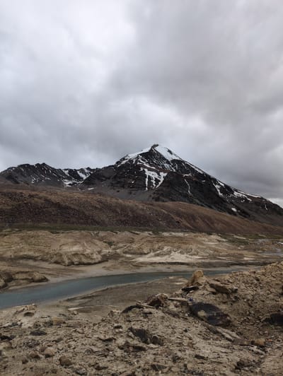 An overcast sky looms over a dramatic mountain landscape. The foreground is a rough terrain of rocks and dry earth, sloping down to a narrow, winding river. Beyond the river, rolling hills of brown and tan earth rise, transitioning into the imposing mountain range. The mountains are rugged, with dark rock faces interspersed with patches of snow, particularly on the highest peak. The overall scene is one of stark, natural beauty, devoid of any human presence or man-made structures. The location is identified as Killingsarai, India.