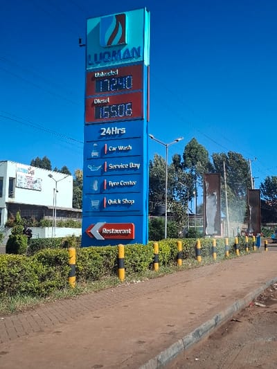 The image shows a gas station sign with a digital display showing the price of unleaded and diesel fuel. The sign is located on the side of a paved road in Nduma, Kenya, at the coordinates -1.157, 36.639. The sign is blue and red, with the words "Luqman" at the top. There are also a number of services listed on the sign, including car wash, service bay, Tyre center, Quick Shop, and Restaurant. The sign is surrounded by a row of yellow and black posts, and there are trees and bushes in the background. There are no people visible in the image.
