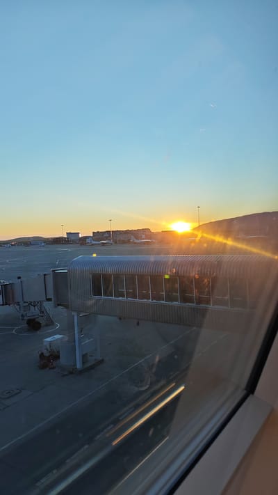 The image captures a sunrise at an airport in Ugolnye Kopi, Russia. The sun is low on the horizon, casting a warm, golden glow across the scene. An airport jet bridge, a covered walkway connecting an airport terminal to an aircraft, is prominently featured in the foreground, extending across the tarmac. Several aircraft are visible parked on the tarmac, along with airport infrastructure such as buildings and light poles. The sky is clear and a soft blue, transitioning to warmer hues near the horizon. The overall impression is one of a calm, early morning at an airport, bathed in the gentle light of the rising sun.