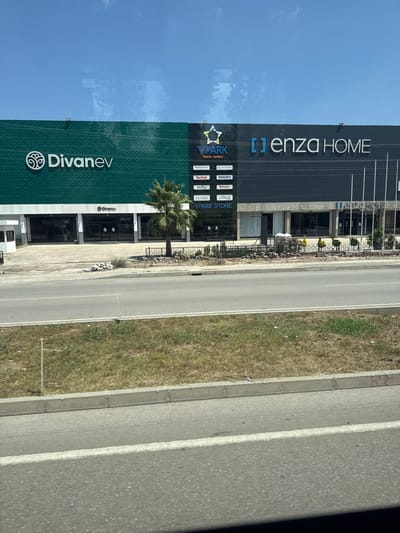 In Osmaniye, Turkey, a commercial building complex is visible from a roadside perspective. The building is divided into two distinct sections. The left section is a vibrant teal color with the branding "Divanev" prominently displayed, accompanied by a circular logo. The right section is dark gray and features the branding "[ ] enza HOME" in large white lettering. Below "enza HOME," there is a list of smaller brand names, including "VPark" and "VPark Store," along with various product brands. A palm tree stands in front of the building, near the entrance to the "Divanev" section. The building complex is situated across a road from a grassy median and embankment. The sky above is clear and blue, with some reflections visible on a glass surface, likely from the vantage point of the photographer.