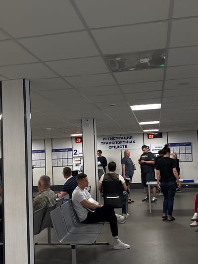 This is a medium shot, eye-level, indoor photograph taken at the State Automobile Inspectorate (GAI) office located in Pargolovo, Russia. The scene depicts a waiting area with several people. In the foreground, on the left, a man in a camouflage shirt is seated on a bench. To his right, a man in a white t-shirt and black pants is also seated. In the background, a digital display shows "02", indicating a queue number or window. Further back, a large banner reads "РЕГИСТРАЦИЯ ТРАНСПОРТНЫХ СРЕДСТВ" (Registration of Vehicles). Another digital display shows "03". Several individuals are standing and waiting in line or at counters. Information boards are mounted on the walls. The environment appears to be a functional, official building with tiled ceilings and fluorescent lighting. The overall atmosphere is one of public service administration.
