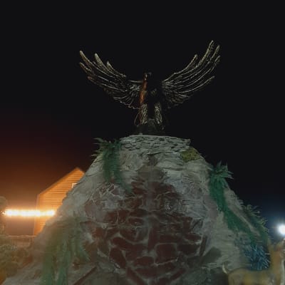 The image captures a nighttime scene featuring a statue of an eagle with outstretched wings, positioned atop a rock formation. To the left of the rock formation is a wooden building with a sloped roof, illuminated by string lights. The rock formation itself is textured and appears to be adorned with artificial greenery. A portion of a deer statue is visible in the lower right corner of the frame. The background is dark, indicating it is nighttime. The scene is located in the city of Sosnovka, Kyrgyzstan.