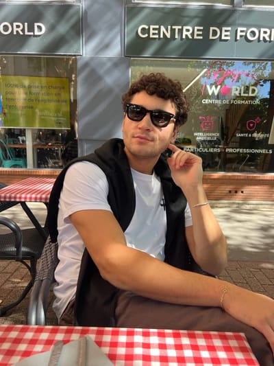 The image displays a young adult male seated at an outdoor café table in Orléans, France. The background identifies the location as "WORLD CENTRE DE FORMATION," situated at 7 Rue Charles Sanglier, 45000 Orléans.
The subject, with dark, curly hair, wears black wayfarer-style sunglasses, a white crew-neck t-shirt, and brown trousers, with a black hoodie draped over his shoulders. A silver-colored bracelet is on his left wrist, and a delicate gold-colored chain bracelet with a small charm is on his right forearm. He is positioned facing slightly to the viewer's left, with his left hand raised, fingers touching his right jawline/ear area. His expression is composed with a faint smile.
He is seated at a table covered with a red and white checkered tablecloth, upon which a white folded napkin and visible cutlery (fork and knife) are placed in the foreground. The chair is a dark, woven-style outdoor model. The background features a building facade with large glass windows. Above the windows, dark grey panels display white signage reading "WORLD" and "CENTRE DE FORMATION." Window decals in pink script repeat "WORLD CENTRE DE FORMATION," while green text advertises "100% de prise en charge pour une formation..." Additional icons and text denote "INTELLIGENCE ARTIFICIELLE" and "SANTÉ & SÉCURITÉ." The interior of the building is partially visible through the reflective glass. The scene is brightly lit by direct sunlight, casting distinct shadows on the building and the man's right arm, indicating an outdoor urban setting on a clear day.
