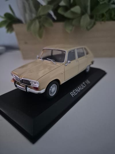 A die-cast model of a beige Renault 18 is displayed on a black plinth. The model car is positioned at an angle, showcasing its front and side profile. The plinth has the words "RENAULT 18" printed in white text. In the blurred background, a planter with green foliage is visible. The overall scene is well-lit, with the focus sharp on the model car. There are no people or any other identifiable objects in the image. The image has no discernible relation to Iskrets, Bulgaria.