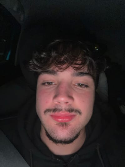 A dimly lit selfie features a young man with curly dark hair and light eyes. He has a mustache and a short beard. The camera angle is slightly skewed, with his face tilted to one side. He wears a dark hoodie, mostly obscured by shadows. The background is dark, suggesting an indoor or nighttime setting. The image has a casual, unfiltered quality.