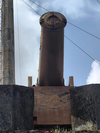 An outdoor scene in Phú Xuân Ward, Vietnam, features a large, heavily corroded artillery piece. The primary subject is a substantial, rust-colored cannon barrel, oriented upwards and slightly right, exhibiting extensive surface degradation and flaking at its muzzle. It is secured on a rectangular, rust-toned metal carriage that displays an embossed dragon motif on its visible front panel, also showing significant corrosion. In the immediate foreground, a dark, rough-textured concrete structure, likely a defensive wall or pedestal, partially obstructs the cannon's base. Sparse vegetation grows along the top edge and base of this concrete structure. To the left, a tall, octagonal concrete support column rises vertically, showing weathering. The upper background consists of a clear to partly cloudy blue sky, intersected by several dark overhead utility lines. The scene is viewed from a low-angle perspective.