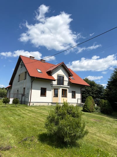 This is an eye-level, medium shot of a two-story house with a red tile roof, located in Strzyżowice, Poland. The house is painted white with a yellow accent on the lower portion of the front facade. It features several brown-framed windows, a dormer window on the roof, and a balcony with a black railing on the second floor. A black power line runs diagonally across the upper portion of the frame.
The house is situated on a sloped, green lawn that is neatly mowed, with visible mowing patterns. There are several evergreen bushes and small trees planted in the yard. In the background, there is a dense line of dark green trees. The sky above is a clear blue with scattered, fluffy white clouds. The overall scene depicts a residential property on a sunny day.
