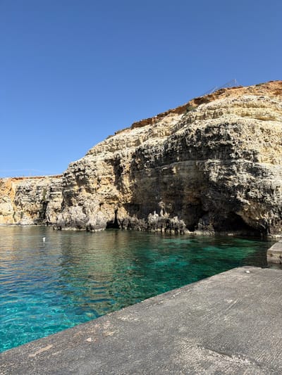 This image captures a vibrant coastal scene, characteristic of Mellieħa, Malta. The primary features are a dramatic rock cliff, clear blue-green waters, and a concrete platform, all bathed under a bright, cloudless sky.
The left side of the frame is dominated by a towering, rugged cliff face composed of stratified, light-colored rock with darker, eroded sections and small caves or overhangs at its base. Hints of sparse vegetation are visible along the cliff's upper edge. Near the top of the cliff, a light-colored metallic frame structure, possibly a safety railing or support, can be seen.
The water, which occupies the mid-right portion, is exceptionally clear and exhibits striking turquoise and emerald hues, indicating shallow depths and pristine conditions. Sunlight creates intricate ripple patterns on its surface, reflecting the surrounding rock and sky.
On the far right, a grey, textured concrete platform or walkway forms a sharp diagonal edge, bordering the clear water. A small, light-colored object, possibly a piece of debris or a shell, rests on its surface.
The sky, a deep and unblemished blue, fills the upper left area, signifying a sunny day with excellent visibility. The overall lighting suggests midday or early afternoon. No people or activities are visible, contributing to a sense of natural tranquility and undisturbed beauty. There is no visible text in the image.