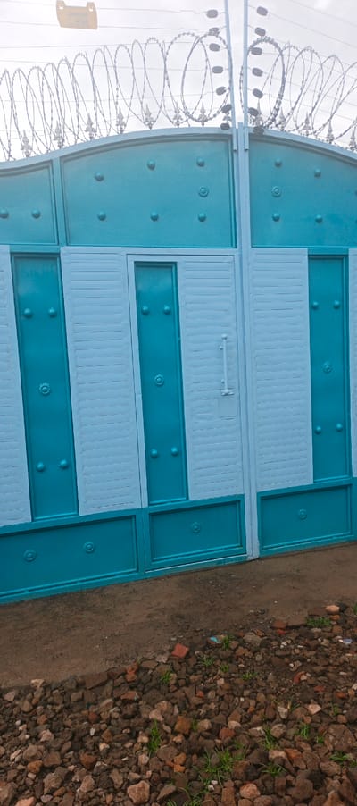 A two-tone blue metal gate stands on rocky ground. The top of the gate is adorned with decorative points and barbed wire. The gate's design alternates between solid blue panels with raised circular embellishments and sections of light blue horizontal slats. A simple white handle is visible on one of the central gate panels. The sky is overcast, creating a muted light on the gate.
