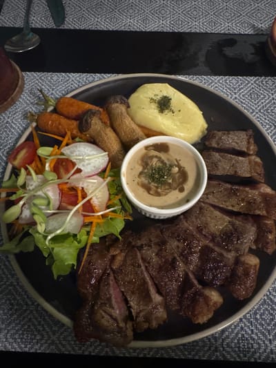 A dark, circular dinner plate is centered, presenting a prepared meal in a top-down, close-up view, consistent with a dining establishment in Ban Ko Li Pe, Thailand. The plate holds a sliced, grilled steak, appearing medium-rare to medium, distributed across the right and lower right portions. A small, white, fluted ramekin in the center contains a creamy, light brown sauce, possibly mushroom gravy, garnished with chopped green herbs. Adjacent to the sauce, in the upper right quadrant, is a portion of smooth, pale yellow mashed potatoes, topped with dried green herbs. The upper left of the plate contains several whole, roasted orange carrots and two large, elongated, sautéed mushrooms, light brown in color. A fresh green salad occupies the mid-left, consisting of green lettuce, sliced red cherry tomatoes, thin strips of orange carrot, and thin slices of white radish, topped with green sprouts. The plate rests on a placemat featuring a repeating blue and white geometric pattern. A dark, possibly black, rectangular table runner is visible horizontally across the upper background. A metallic utensil, likely a spoon, and the rim of a dark-colored cup or glass are partially visible in the upper left background. No individuals or active interactions are depicted.