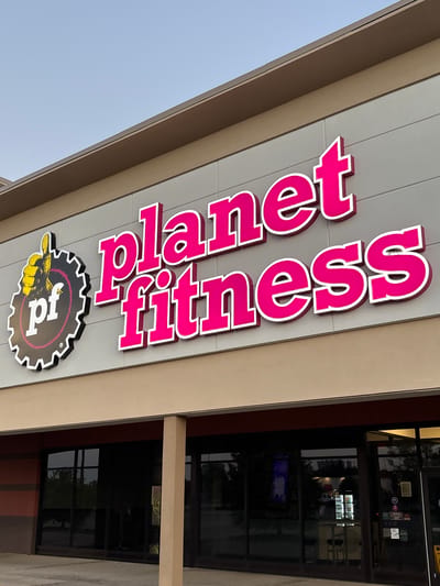 The image shows the exterior of a Planet Fitness gym. The sign for Planet Fitness is prominently displayed in large, bright pink letters with white trim, along with the Planet Fitness logo featuring a thumbs-up inside a gear. The building has a light-colored facade with a dark brown trim along the bottom, and large glass windows and doors are visible. The sky is a pale blue, suggesting it is either early morning or late afternoon. The gym appears to be located in Mount Vernon, United States of America. There are no people visible in the image.