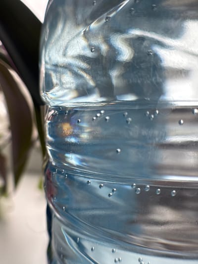 The image shows a close-up view of the lower portion of a clear plastic bottle, likely filled with water. The bottle has a ribbed or contoured design, with numerous small air bubbles clinging to the inner surface. The plastic is slightly distorted, creating a wavy effect and refracting light. The bottom of the bottle rests on a dark, slightly blurred surface, possibly a table or stand. In the background, there is a blurry glimpse of vegetation. There is no indication of location or inhabitants.