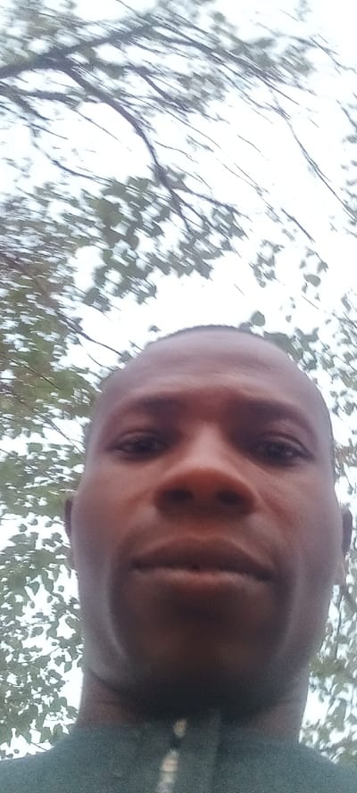 A close-up selfie reveals a man with dark skin, a shaved head, and intense, dark eyes. The camera's low angle emphasizes his nose and jawline. Behind him, blurry tree branches and leaves create a diffused, light backdrop, suggesting an outdoor setting. The lighting is soft, and the color palette is subdued, dominated by browns, greens, and whites. He is wearing what appears to be a dark garment.