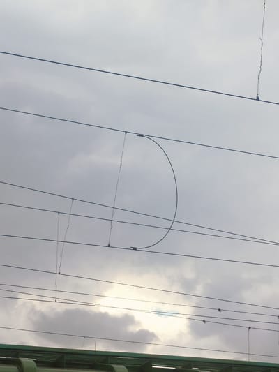 The image captures a section of overhead power lines against a cloudy sky. The lines are taut and suspended at various heights, with some showing attachments and clips. Notably, a semicircular loop formed by a wire is visible in the mid-section of the frame, suspended between two horizontal wires. In the lower part of the image, a green structure, possibly part of a building or infrastructure, is partially visible. The sky is overcast with areas of brighter light breaking through the clouds, suggesting a daytime scene. The location is identified as Salsk, Russia.