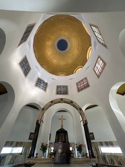 This is an interior shot of a domed building, likely a church or chapel, in the Emek HaYarden Regional Council, Israel. The focal point is a large, ornate golden dome that dominates the upper portion of the image, with a dark circular opening at its center. The dome is surrounded by a white, curved ceiling structure with several recessed rectangular panels. These panels contain stained glass with Latin inscriptions, possibly religious verses or names, as indicated by the readable text like "BEATI MITES," "SPIRITV," and "BEATI PACIFICI." The overall architecture suggests a place of worship, possibly with a Byzantine or modern interpretation of traditional religious design.

To the right, an altar area is visible, featuring a dark, tiered structure topped with a cross. The altar is adorned with flowers in vases and what appears to be a candle. The background of this area is marble. The lighting suggests it could be daytime, with natural light entering through a rectangular window visible in the upper right. The mood is solemn and reverent, characteristic of a religious setting. The image is captured from a low angle, looking upwards towards the dome and ceiling.