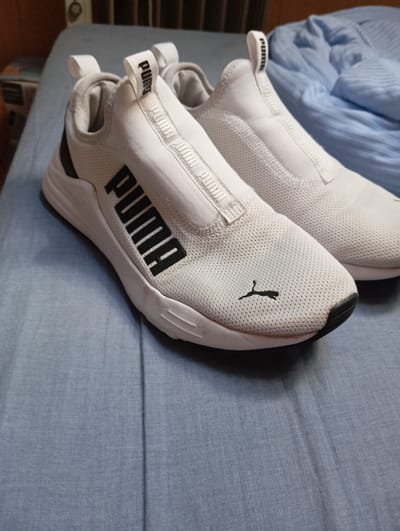 The image displays a close-up view of a pair of white Puma athletic shoes placed on a blue fabric surface. The shoes are positioned diagonally, with the left shoe slightly forward and overlapping the right shoe. Both shoes are slip-on style, featuring a white mesh upper with a black Puma logo prominently displayed on the side of the left shoe. The right shoe also has a black Puma logo visible on its toe. A black strip runs along the heel of both shoes. A zipper detail is present on the front of the shoes, above the laces. The background consists of blue fabric, possibly a bed or couch, with some rumpled areas. The overall lighting suggests an indoor setting. This scene does not depict any specific location in the city of Parchim, Germany.