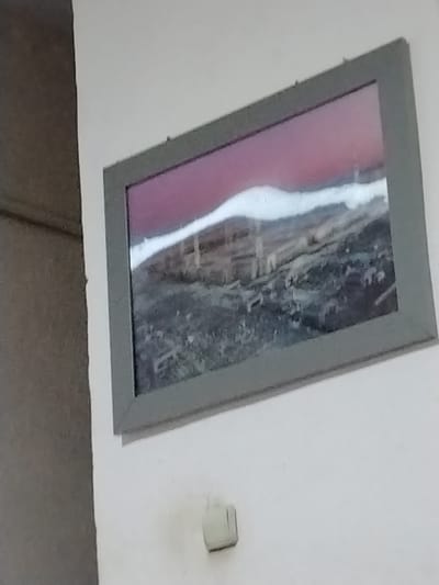 A framed painting depicting a landscape with a purple sky and a white wavy band across a dark, textured ground is hung on a white wall. Below the painting, a small, light-colored electrical outlet is visible on the wall. The scene appears to be indoors, possibly in an art gallery or a home. The provided image does not contain information about the city of Nukus, Uzbekistan.