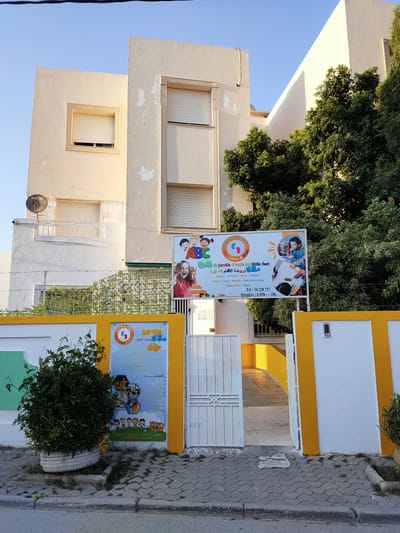 The image depicts the exterior of a building with a sign for "Little Feet" kindergarten. The building appears to be in a residential or commercial area. The sign is brightly colored and features cartoonish images of children and a school bus. It also lists various activities offered, such as English, French, music, theater, Quran, calculations, mental development, and sports. The building has windows with shutters, and there is a satellite dish mounted on the wall. Trees and greenery surround the building, and a walkway leads to an open gate. The overall impression is that of a well-maintained educational facility. The location is identified as Unknown, Tunisia.