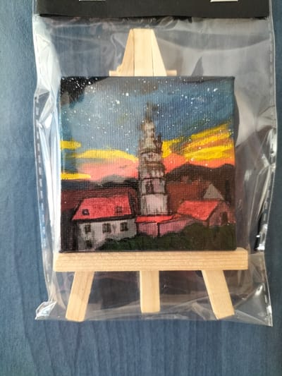 This media file shows a miniature acrylic painting of a cityscape, likely in České Budějovice, Czechia, displayed on a small wooden easel. The painting depicts a clock tower, resembling the Black Tower (Černá věž) in České Budějovice, with several red-roofed buildings in the foreground. The scene is set at dusk or dawn, with a colorful sky featuring shades of dark blue, orange, yellow, and pink. Tiny white dots scattered across the sky suggest stars or perhaps distant lights. The painting is enclosed in clear plastic packaging, attached to a black header. The wooden easel has three legs, two in the front and one supporting the back, made from unfinished light-colored wood. The background of the image is a textured blue fabric.
