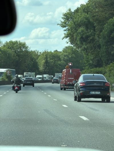The image captures a view from a vehicle traveling on a highway in Savigny-sur-Orge, France, indicated by the speed limit sign showing "110 RAPPEL." The highway is lined with lush green trees on both sides. In the foreground, a motorcycle with a rider wearing a leather jacket and helmet is moving away from the camera. The motorcycle has a license plate that is partially obscured but appears to read "53 KW." Ahead of the motorcycle, several vehicles are visible, including a dark-colored sedan, a red truck with "CLOXAM" written on it, and multiple vans and trucks. The sky is partly cloudy with a bright, hazy sun. The road surface is asphalt, marked with white lane lines. The overall scene depicts a typical highway traffic situation on a clear day.