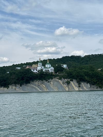 An eye-level, medium shot captures a serene body of water in the foreground, with a rocky cliff and a church nestled on a lush green hillside in the background. The water, a muted blue-green, displays a gentle ripple pattern, suggesting a calm day. The cliff face is a striking geological formation, characterized by layered sedimentary rock in shades of gray, beige, and white, with patches of greenery clinging to its surface.
Above the cliff, a verdant forest covers the hillside, dotted with several buildings. The most prominent structure is a white church with green domed roofs, featuring golden crosses. The church appears to be part of a monastery or religious complex, with other smaller buildings also visible. The architecture suggests a traditional Russian Orthodox style. A few modern houses with clean lines and large windows are also interspersed among the trees.
The sky is a mix of soft blue and scattered white clouds, some of which are fluffy and illuminated, while others are more wispy and elongated, indicating a partly cloudy day. The overall scene evokes a sense of tranquility and natural beauty, with the man-made structures harmoniously integrated into the landscape. This scene is likely the Monastery of the Assumption of the Mother of God, located on the shores of Abrau Lake in Abrau-Dyurso, Russia.