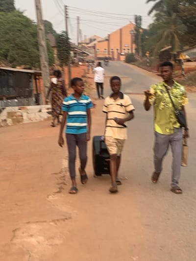 Three men stand in a line on a road. The first man wears a green and yellow patterned shirt with grey pants and sandals. The second wears a tan and white striped shirt with khaki shorts, standing beside a black suitcase. The third has on a blue and white striped shirt, grey pants, and sandals. A white-shirted person walks further down the street. Buildings, trees, and power lines are visible in the background.