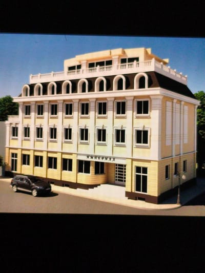 A depiction of the Hotel Orpheus in Devin, Bulgaria. The hotel is a three-story building with a yellow and white facade, featuring arched windows on the top floor and a flat roof with a decorative railing. A dark-colored SUV is parked in front of the building. A street lamp stands near the corner of the building. Trees can be seen in the background under a blue sky.