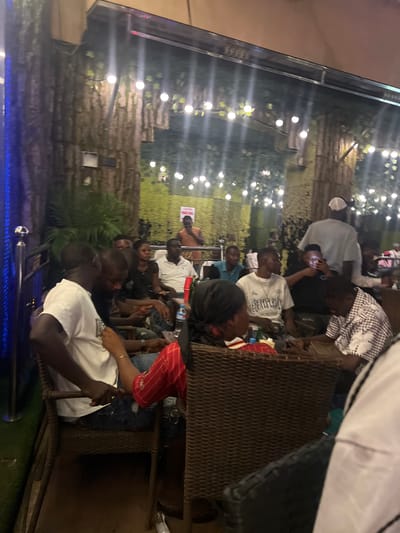 This image captures an outdoor gathering at a venue with a naturalistic, jungle-themed decor in Oba, Nigeria. The background features artificial trees with textured bark, adorned with numerous string lights that cast a warm glow. A green, leafy artificial plant is visible to the left, adding to the lush ambiance. The foreground shows several individuals seated at tables, engaged in conversation and socializing.

In the left foreground, a person wearing a white t-shirt and blue jeans is seated, with their arm resting on a wicker chair. To their right, another individual in a black t-shirt and jeans is partially visible, holding a drink. Further back and to the right, a person in a white polo shirt is seated, and next to them, a person in a black and white checkered shirt is looking down at their phone. The group appears to be relaxed and enjoying the evening. The lighting is dim, with the string lights being the primary source of illumination, creating an intimate and festive atmosphere. The presence of tables and chairs suggests a seating area designed for patrons.