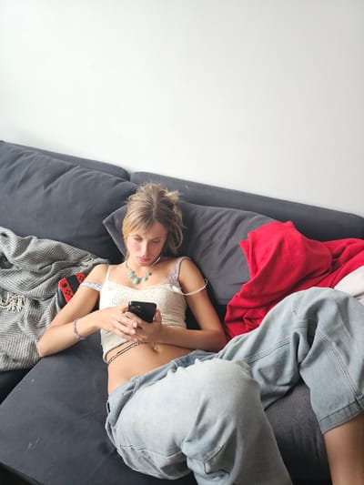In a brightly lit indoor setting, a young woman with blonde hair tied up in a bun is casually lounging on a dark grey couch. She is wearing a white lace crop top, light blue baggy jeans, and a necklace with turquoise stones. Her left arm is adorned with a purple beaded bracelet. She is holding a black smartphone in her hands and appears to be looking at it. Beside her on the couch, to her left, is a grey patterned blanket and a red Nintendo Switch game console. To her right, a vibrant red garment is draped over the couch. The wall behind the couch is a plain white. The scene suggests a relaxed, at-home environment. The location of Pantin, France is not discernible from the image.