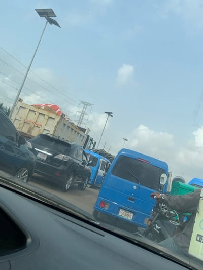 The scene depicts a traffic queue on a multi-lane road in Jakande, Lagos State, Nigeria, viewed from the interior of a vehicle. The sky is light blue with scattered white clouds.
In the immediate foreground, the dashboard and lower windshield of the observer's vehicle are visible. To the right, a person wearing a dark top is seated on a black "GENSTAR" motorcycle, stationary next to a blue Suzuki van. This van, license plate "LSD 648YL", is positioned directly ahead of the motorcycle and features a rear wiper. Another blue van is partially visible behind the Suzuki van.
In the adjacent lane to the left, a black Lexus RX 350 SUV with license plate "50G00LA" (partially obscured) is stopped. Further left, the rear of a dark-colored sedan is visible. Behind the Lexus, a large dump truck (tipper truck) with "Carrillion" branding on its side carries a load of light-colored material, possibly sand or gravel, partially covered by a red and white striped tarp or fabric.
The environment includes multiple tall metallic streetlights equipped with solar panels. In the background, a large metal electricity transmission pylon and overhead power lines are visible. Distant green vegetation is present on the far left. All vehicles appear to be stationary or moving very slowly, indicative of traffic congestion.