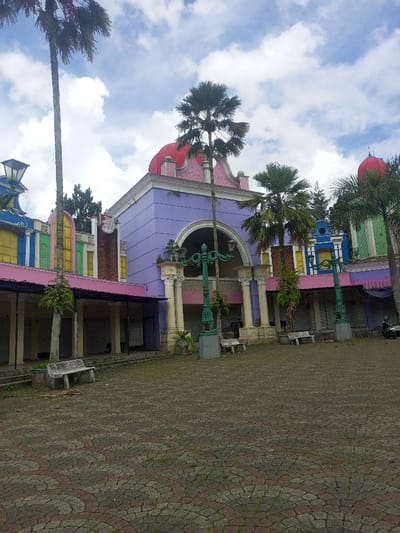 The image depicts a multi-story commercial or recreational building complex located in Cipanas, Indonesia, viewed from a low angle across a paved plaza under a partly cloudy sky. The central structure features a lavender-painted facade with a prominent red dome roof and pink decorative trim. A large arched recess on the second story is flanked by four white fluted columns. Adjacent sections of the complex extend horizontally, displaying an eclectic mix of bright colors including yellow, blue, green, and pink, with various arched openings and lower-level units, many secured by closed roll-down metal shutters.
In the foreground, a plaza is paved with interlocking red-brown and grey/white pavers arranged in a circular pattern, with sporadic weeds and grass growing between them. Two weathered, light-colored stone benches are present on the plaza. Two ornate, tall green lampposts, each with multiple luminaires, stand in front of the building's columns. Several tall palm trees are distributed throughout the scene, some reaching above the roofline. Smaller green foliage is visible near the building's base. No individuals are visible. A dark-colored motorcycle is partially visible parked near the right side of the building complex. The scene appears unoccupied and shows signs of reduced activity.