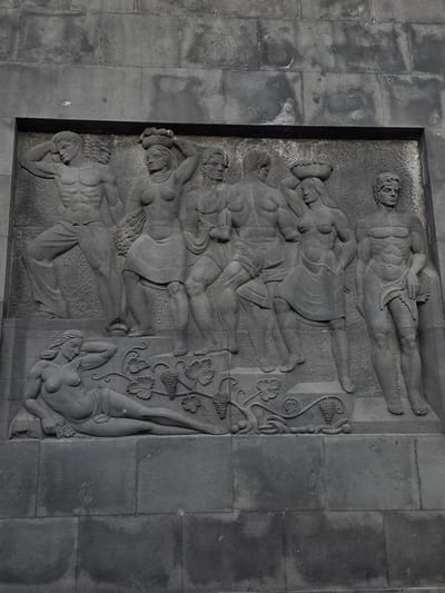 This is a high-angle, close-up, exterior shot of a stone bas-relief depicting a horizontal arrangement of figures and grapevines. The relief is set into a wall made of large, dark gray, rectangular stone blocks.
The main subjects are six human figures, appearing to be men and women, rendered in a stylized, classical manner. They are depicted in various reclining and entwined poses, suggesting a scene of revelry or perhaps a mythological narrative. Some figures are partially draped, while others are more exposed, with details of musculature and anatomy evident. One figure at the top appears to be holding a segmented object, possibly a weapon or tool. Towards the right side of the relief, a cluster of grapevines with leaves and bunches of grapes are intricately carved, entwining with some of the figures. A seventh, smaller figure, appears at the bottom right, seemingly upside down and intertwined with the vines.
The activity suggests a scene of leisure, festivity, or perhaps a struggle or union, amplified by the intertwined nature of the figures and the presence of the grapevines, which often symbolize fertility and abundance. The stonework is detailed, with the carvings showing depth and texture. The lighting suggests it is daytime, with shadows indicating the sun's direction, though the exact time of day or weather conditions are not clearly discernible. The location cues, such as the architectural style of the stone wall and the subject matter, could be indicative of a public monument or a historical building in Santa Cruz de Tenerife, Spain. No text is visible in the image.