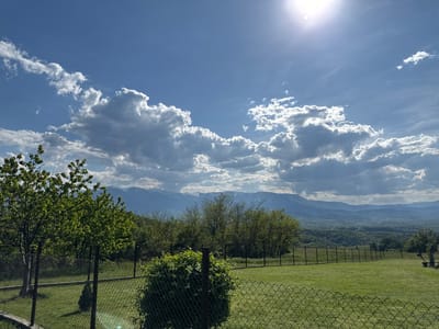 The image depicts a view from a fenced yard in Bokilovtsi, Bulgaria, on a sunny day with a few clouds. A green lawn occupies the foreground, enclosed by a chain-link fence. Beyond the fence, there are several trees and bushes. In the background, rolling hills and mountains are visible, some with patches of snow. The sky is blue with scattered white clouds and the sun shining brightly. There are no people visible in the frame.