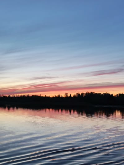 The image captures a serene waterfront scene at twilight. The sky transitions from a deep blue at the top to hues of pink and orange along the horizon, where thin cloud streaks run horizontally. Silhouetted trees line the horizon, their dark shapes reflected in the calm water below. The water is textured with gentle ripples that create a distorted reflection of the sky and trees. The scene appears to be a natural landscape, with no visible human presence or built structures beyond the treeline, suggesting a rural or undeveloped area. There is no information to place this scene in Tarkanovo, Russia.