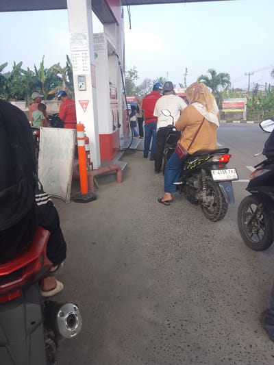 The image depicts a busy gas station in Cibarusah, Indonesia, likely on a bright, clear day. Several individuals are present, with most gathered around gas pumps. A person in a red uniform, possibly a gas station attendant, is interacting with a customer. Multiple motorcycles are parked, some with riders waiting, while others appear to be parked and unattended. One motorcycle in the foreground has a license plate reading "F 4198 FKJ 03-30." The gas station features red and white structures, typical of a Pertamina fuel station. The ground is paved with asphalt, and lush green foliage, including banana trees, is visible in the background. The overall scene suggests a typical day at a local gas station.