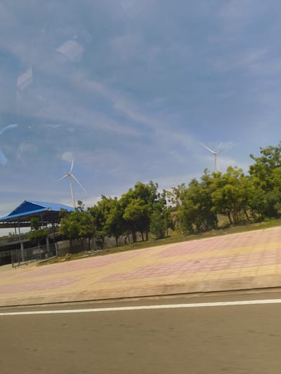 The image depicts a bright, daytime scene featuring two wind turbines against a partly cloudy blue sky. In the foreground, a brown asphalt road is visible with a white line dividing it. Behind the road, a paved walkway with a patterned design of pink and yellow rectangular tiles runs horizontally. Further back, a row of lush green trees forms a backdrop. To the left, a structure with a blue roof and open sides, possibly a shed or a small building, is situated. One wind turbine is positioned behind the structure, while the second is further to the right, partially obscured by the trees. The overall environment suggests an open area, likely in a rural or semi-rural setting. The reflection on the surface in the upper left corner of the image indicates it was taken through a window. The location is identified as Hoa Thang, Vietnam.