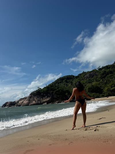 A young woman stands on the shoreline of a beach in Baan Thong Nang, Thailand. She faces the ocean in a black bikini. Green foliage covers a rocky cliff that lines the left side of the beach. The sky is blue with scattered white clouds. Waves wash onto the sandy beach. The light source appears to be from the right.