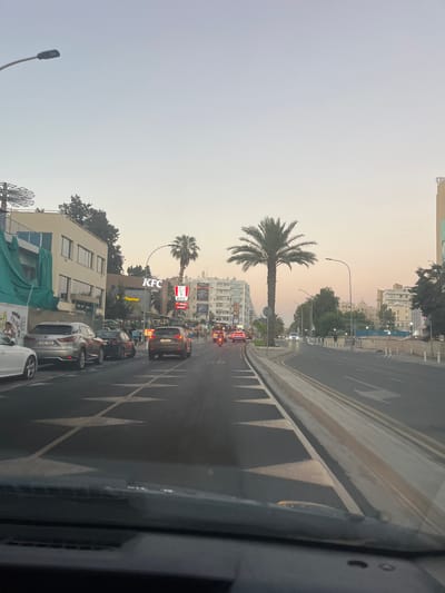 A wide, paved road in the city of Germasogeia Municipality, Cyprus, is captured from a low-angle perspective, likely from inside a vehicle. The road is lined with parked cars and ongoing traffic, including a white SUV, a silver Lexus, a dark sedan, and a dark SUV. The presence of diamond-shaped road markings indicates a transition or warning area. On the left, a multi-story building with a green tarpaulin covering its lower section is visible, adjacent to a KFC restaurant with prominent signage. Palm trees are interspersed throughout the urban landscape, with one particularly large specimen near the center of the frame. Buildings with architectural details typical of Mediterranean cities line the right side of the road, with a pedestrian sidewalk and streetlights. The sky displays a soft, gradient of pastel colors, suggesting either dawn or dusk. The overall scene depicts a typical urban streetscape during a period of moderate activity.