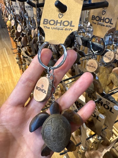 A close-up image shows a person's fair-skinned hand holding a souvenir keychain in what appears to be a retail store or souvenir shop in Panglao, Bohol, Philippines. The keychain consists of a silver-colored metal keyring and a small chain, from which a light brown, circular wooden disc is suspended. This disc has a small heart symbol and the word "BOHOL" etched into its surface. Attached below the wooden disc is a dark brown, carved wooden sea turtle. In the background, a black metal display rack is visible, showcasing numerous other identical or similar keychains. Each keychain on the rack features a brown paper tag with a heart symbol and the text "BOHOL The Unique Gift." Some background keychains also display the "BOHOL" engraved wooden discs and carved wooden figures. The floor beyond the display appears to be light-colored wood.